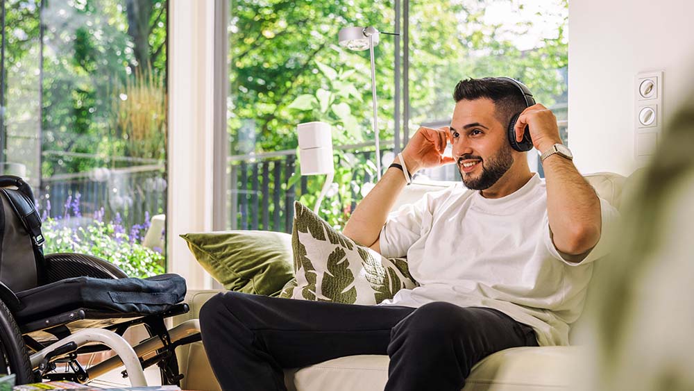 Til høyre sitter en mann på sofaen med hodetelefoner på, han holder hendene på hodetelefonene. Til venstre står en rullestol