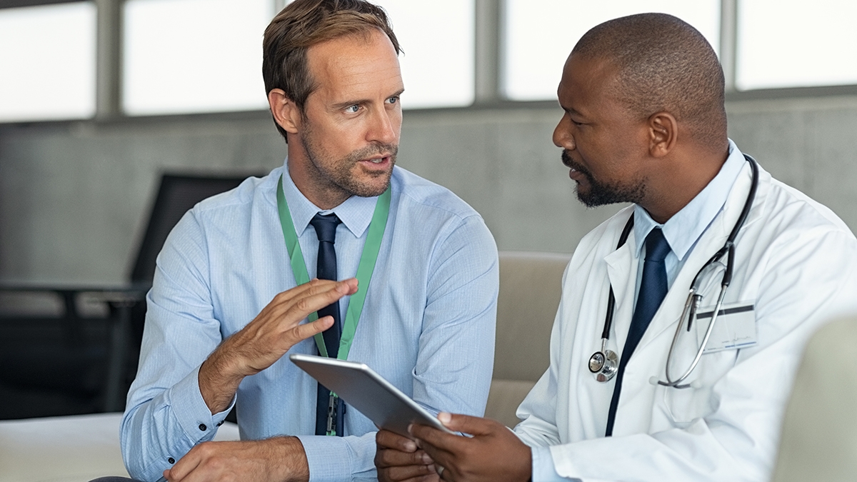 Farmasøytisk representant som snakker med lege som sitter på sofa. Afrikansk moden lege diskuterer resultatene av analysen med spesialist mens han konsulterer diagnosen på et digitalt nettbrett. Lege i samtale med medisinsk rådgiver på sykehusrom som diskuterer pasientrapport.