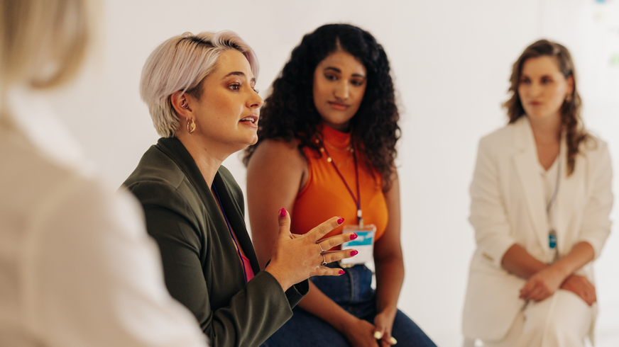 Fire kvinner sitter side om side i Women's Mentoring Program. Målet er å øke synligheten av kvinner i organisasjonen og hjelpe dem med personlig vekst. En annen kvinne observerer den første, en gråhåret kvinne i grønn jakke, mens hun snakker og gir et signal med hånden.  