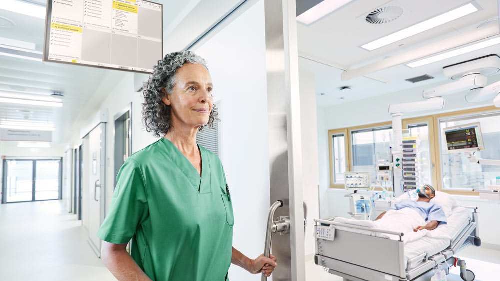 Sykepleier i sykehuskorridoren, utstyrt med silent ICU – i rommet ligger en pasient på intensivavdelingen