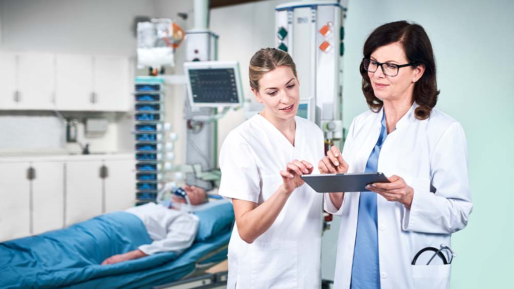 En lege og en sykepleier diskuterer doseringen for B. Braun smart infusion, som administreres direkte fra et nettbrett. Pasienten på intensivavdelingen kan ses i bakgrunnen, koblet til et fullt infusjonsstativ med smarte infusjonspumper.