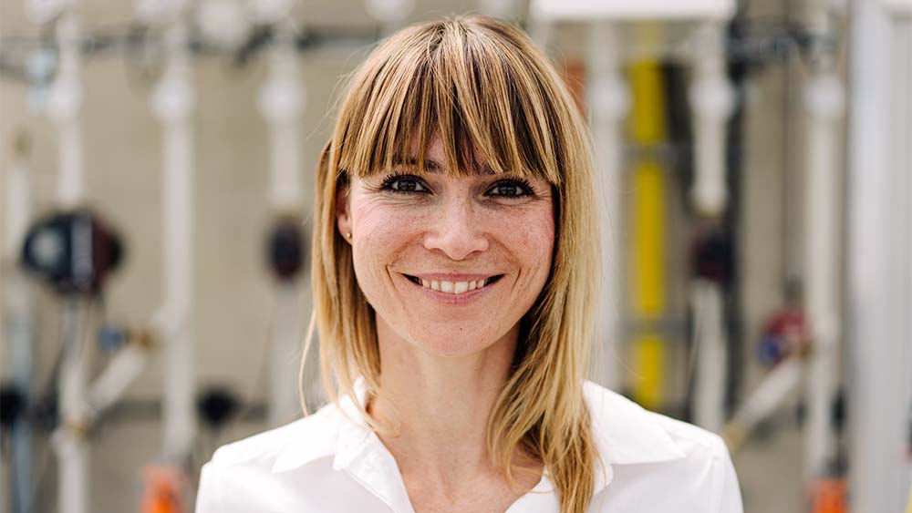 Nærbilde av en kvinne som ser inn i kameraet, smiler og har på seg en hvit skjorte i lyse omgivelser. 