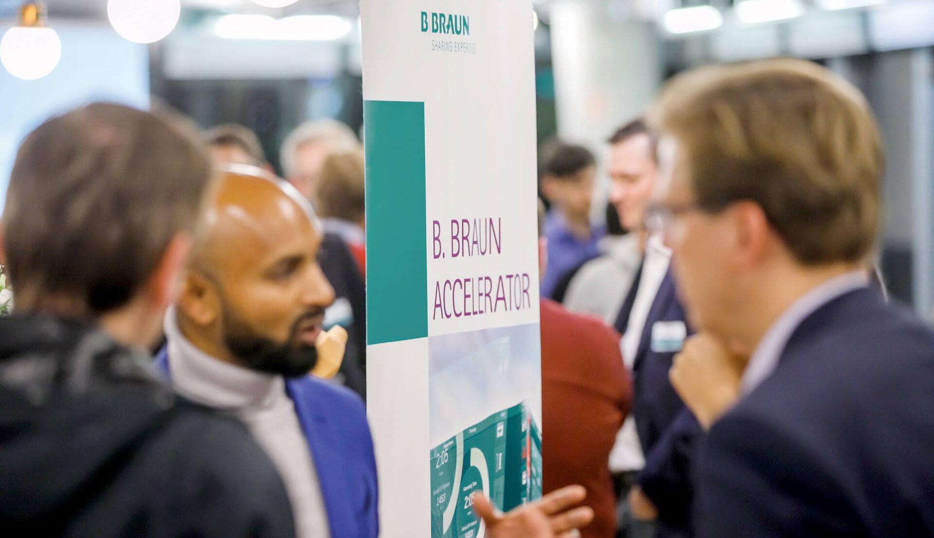 En gruppe mennesker som diskuterer på en konferanse med et banner for B Braun vises i bakgrunnen. 