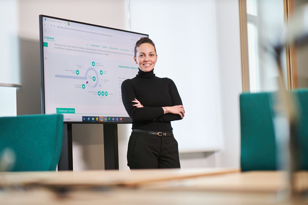 Renata Almeida, en Braun-ansatt, står og smiler foran en TV som viser grafer på et kontor. 